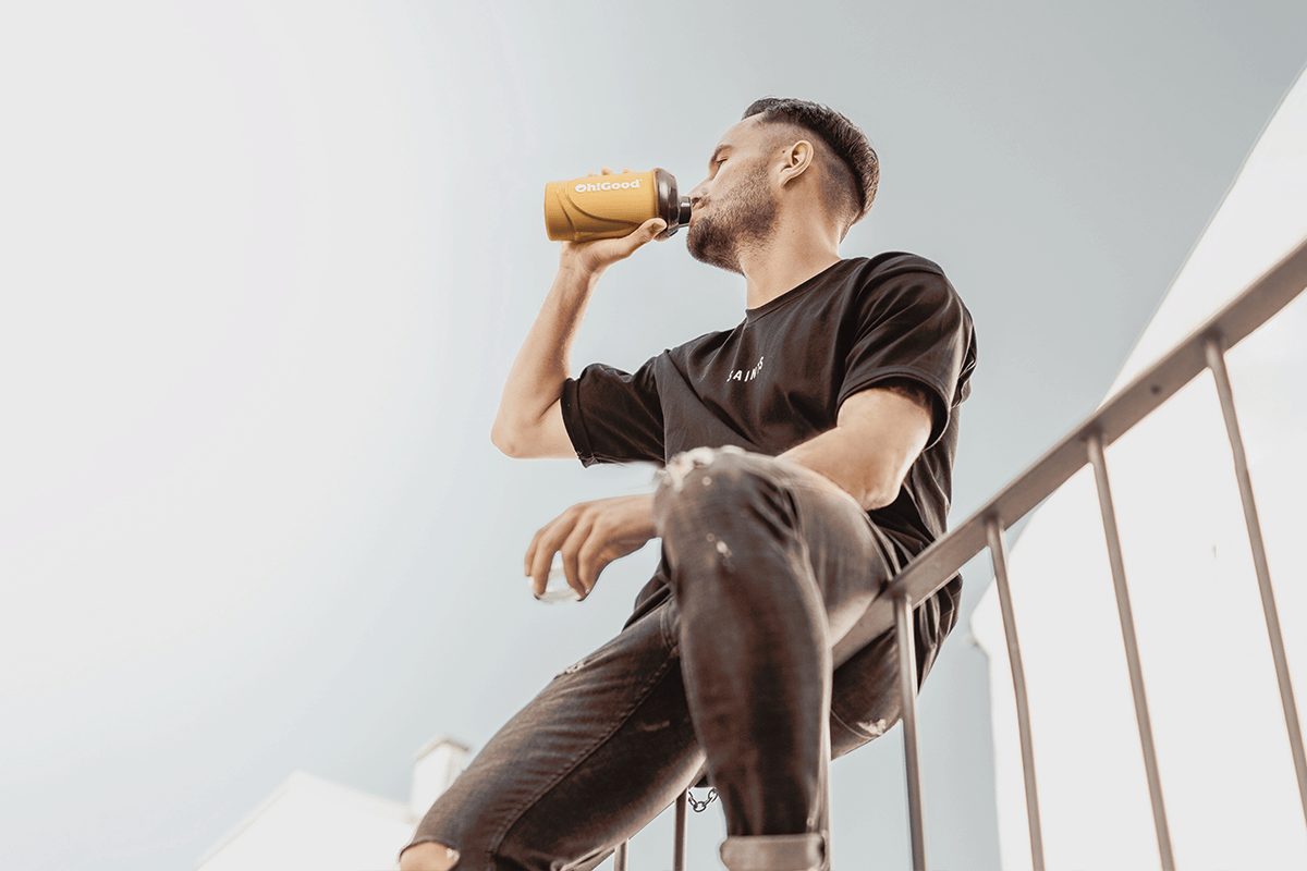 A man sits on a railing, looking up and drinking from an Oh!Good protein shaker.